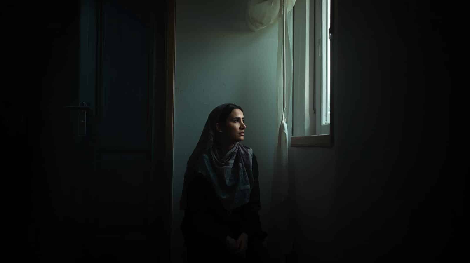 A person sits alone in a dark room, looking out of a window. The mood is quiet and reflective, neither hopeless nor resolved.