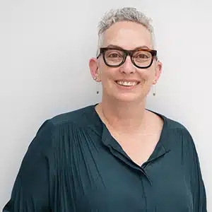 Smiling person with short gray hair and glasses in a blue shirt against a white background. (AI-generated)