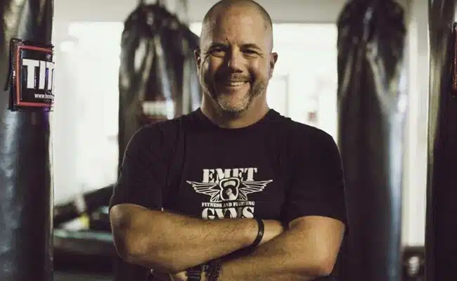Man in gym wearing Emet Gym t-shirt, smiling with crossed arms. (AI-generated)