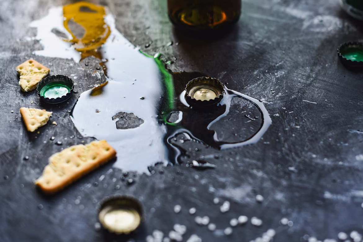 A photograph shows a dirty black tabletop littered with debris including a beer bottle, beer bottle tops, broken pieces of cracker, and a puddle of spilled liquid (presumably beer)
